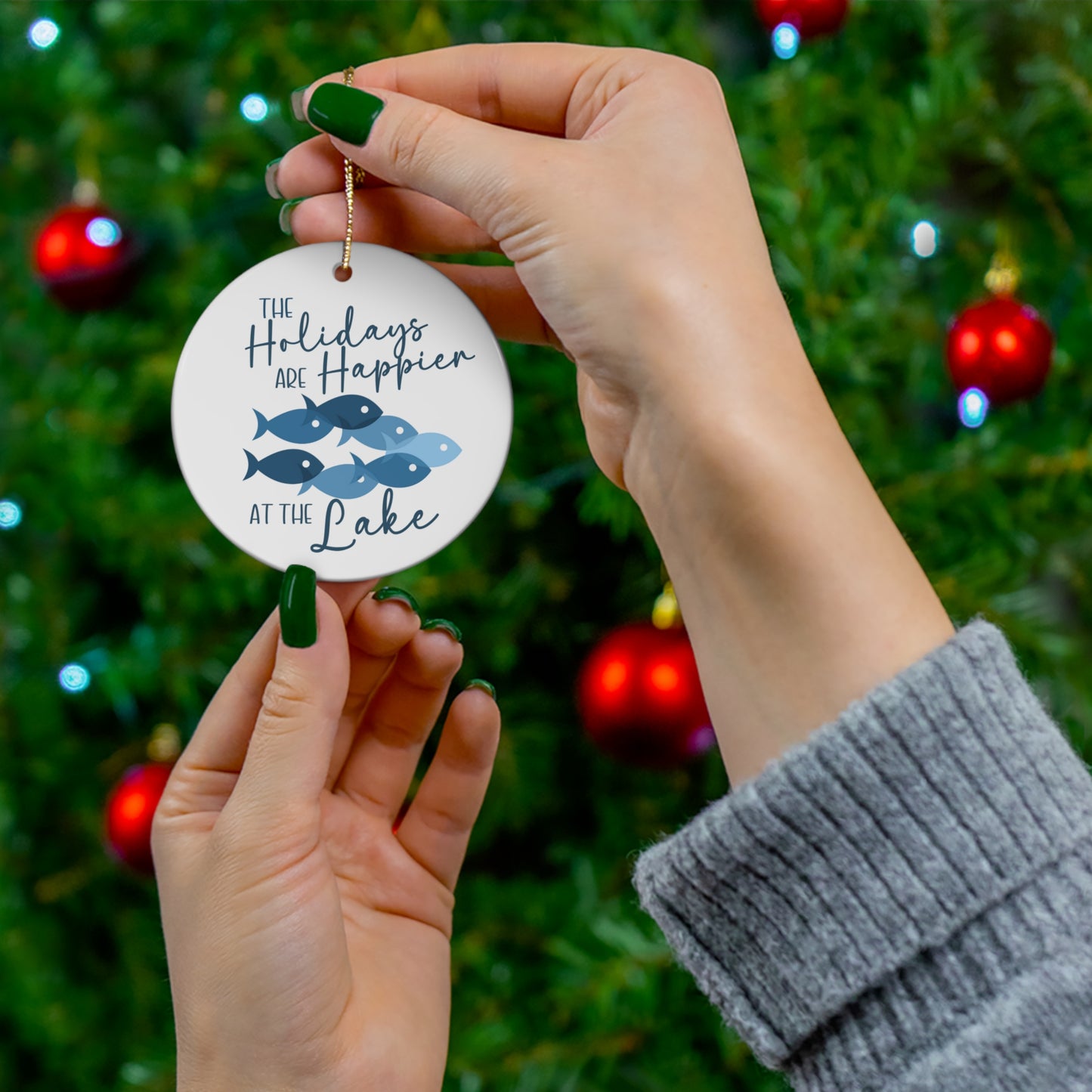 The Holidays are Happier at the Lake Ceramic Ornament, circle shaped tree ornament featuring school of fish and lettering in blue against a white backdrop